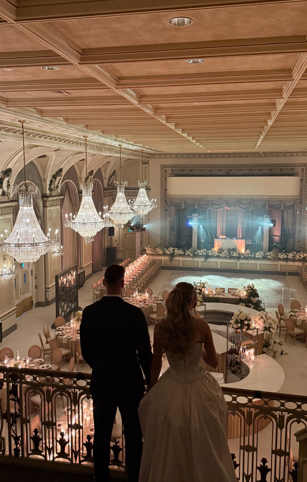 Bride and groom on a balcony overlooking a grand chandelier-lit ballroom set for their destination wedding reception