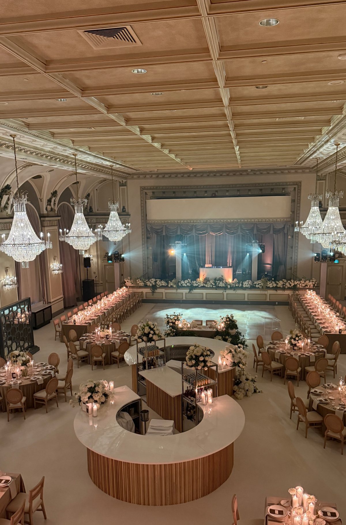Historic ballroom under crystal chandeliers, set with long candlelit tables and a central bar before a destination wedding