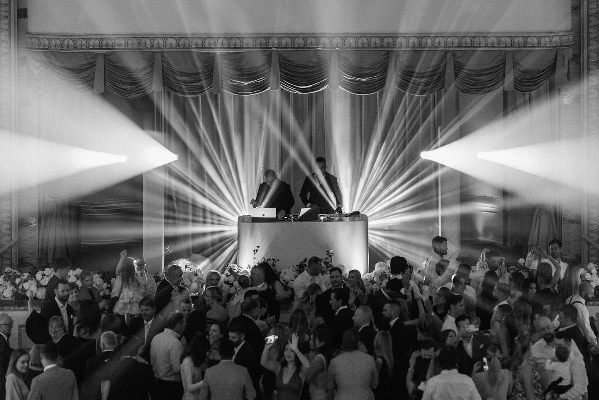 Light beams fanning out from the booth across a packed dance floor in a grand draped ballroom, guests dancing shoulder to shoulder