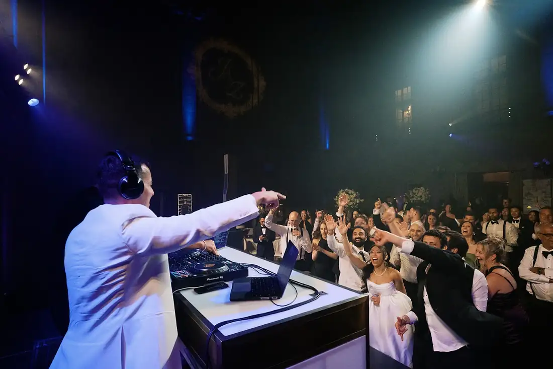 DJ MAJD facing a packed wedding dance floor, arms outstretched as the bride, groom and every guest cheer back
